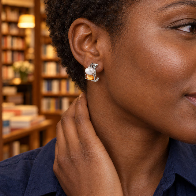 A woman with short coiled hair wears a silver-tone pearl and citrine earring beside warmly lit bookshelves.