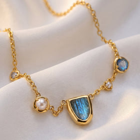 The gold-tone necklace lies on softly folded fabric with its blue stone and white pearls in view.