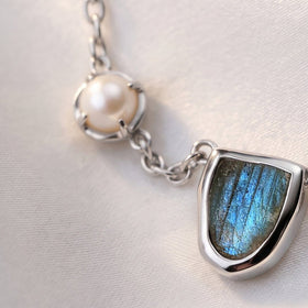 A round white pearl sits in a silver-tone prong setting beside linked chain and part of the blue centerpiece.