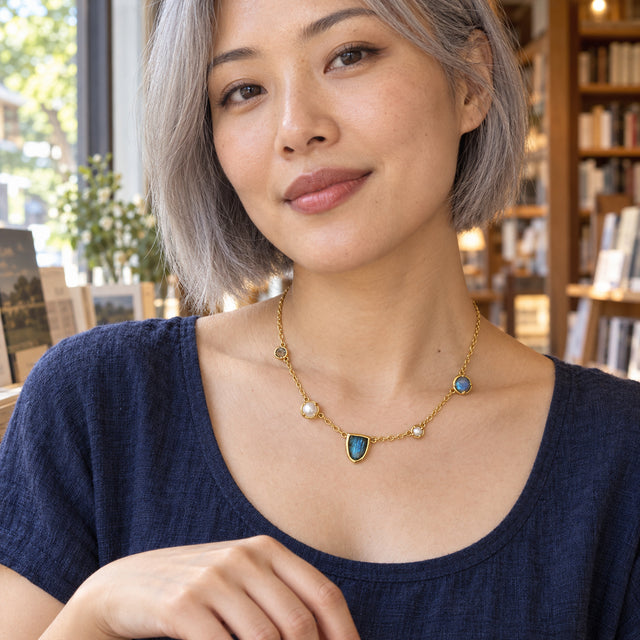A woman with a short gray bob wears the gold-tone labradorite and pearl necklace with a navy top beside a bookshop window.