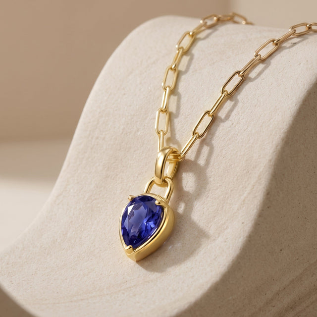 An angled view shows the yellow-gold-tone pendant setting and blue gemstone against a pale textured stone.