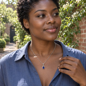 A woman in a slate-blue shirt wears the silver-tone blue pendant necklace in a leafy brick courtyard.