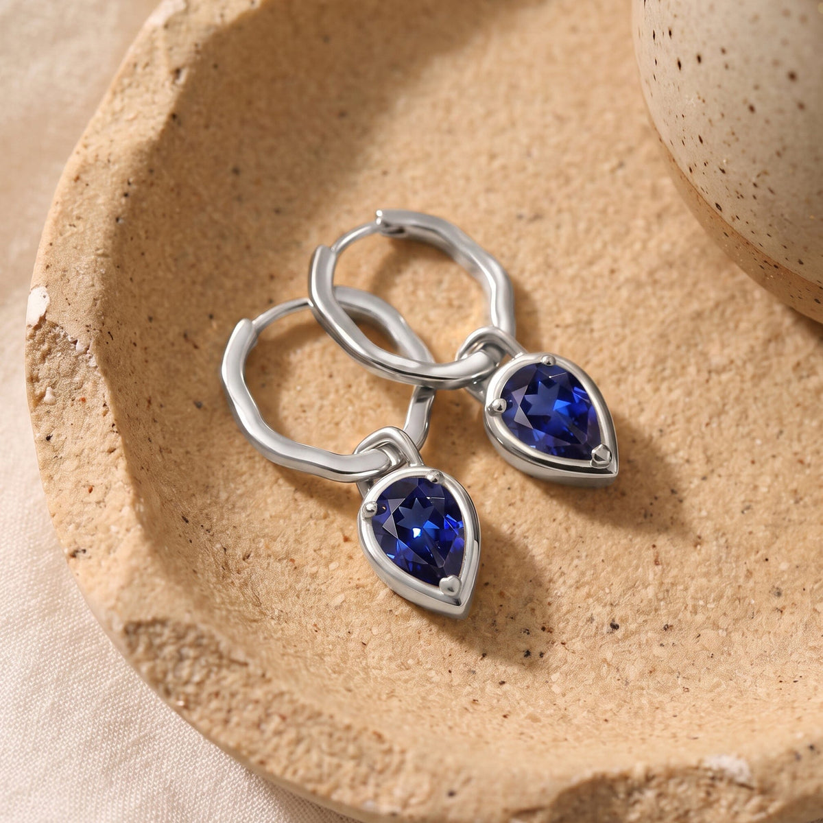 Silver-tone sapphire hoops lie together on a textured beige stone tray.