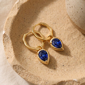 Yellow gold-tone hoop earrings with faceted blue drops are arranged on a speckled ceramic tray.