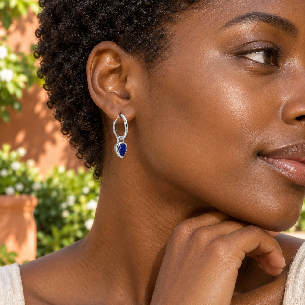 A woman with short curls wears a silver-tone blue sapphire hoop beside sunlit foliage and a terracotta wall.
