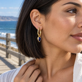 A woman with a dark bob wears a yellow gold-tone sapphire hoop on a coastal boardwalk with the sea behind her.
