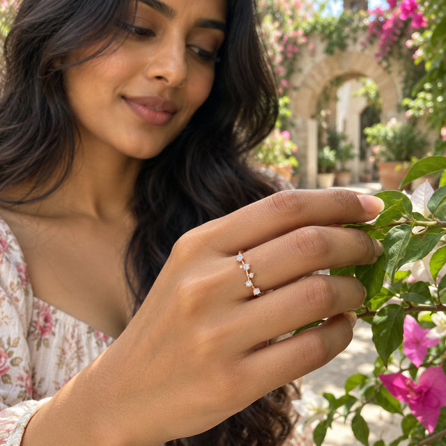 A rose gold vermeil ring with pale round stones is worn on a hand touching green leaves in a flowering courtyard.