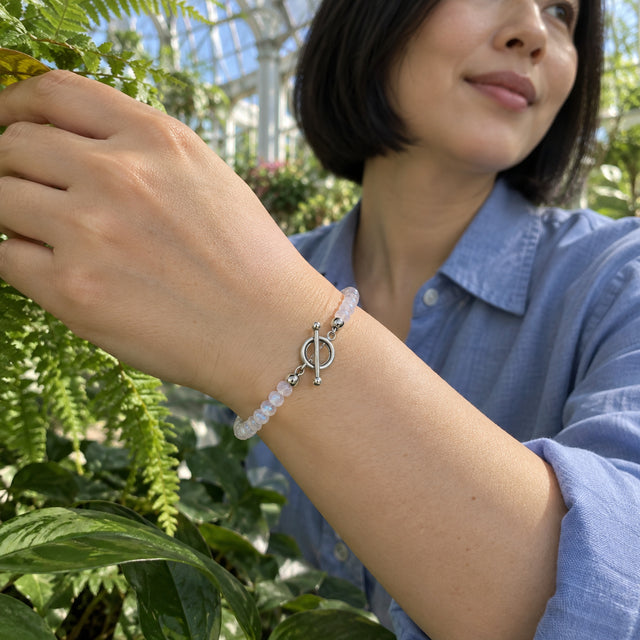 A silver clasp and pale moonstone beads circle a woman's wrist beside fern leaves in a glass conservatory.