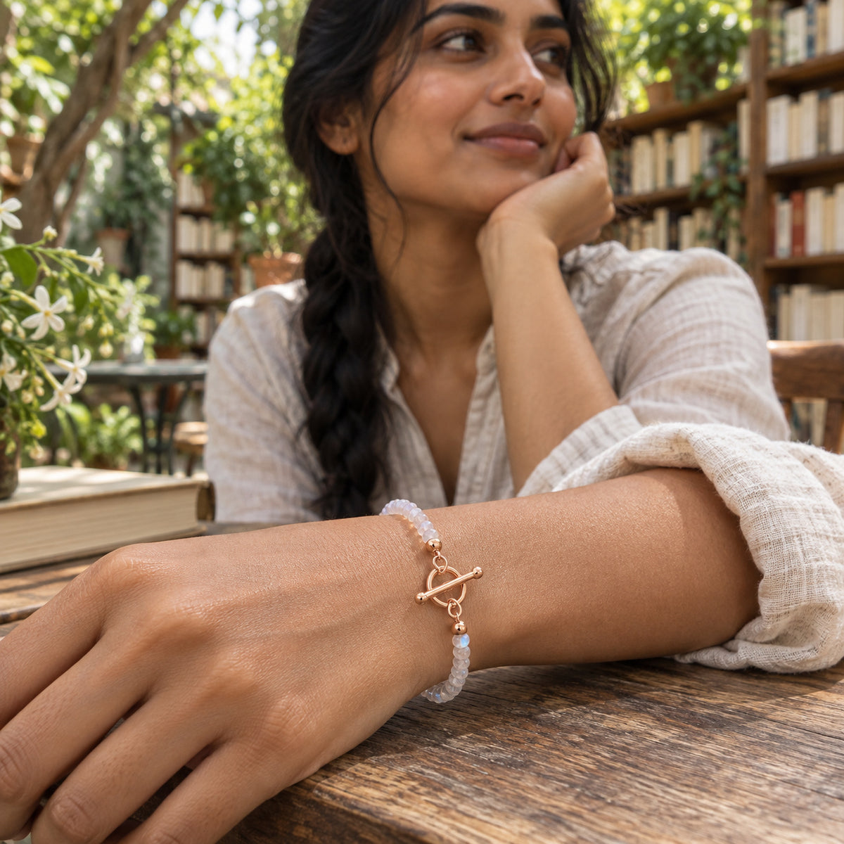 A rose gold clasp joins moonstone beads around a woman's wrist resting on a wooden table in a leafy bookshop.