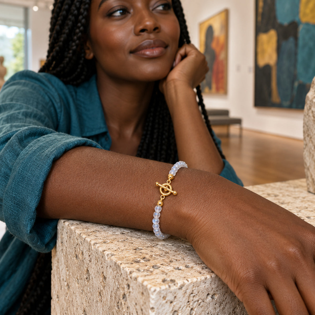 A yellow gold toggle clasp and pale moonstone beads encircle a woman's wrist resting on a stone console in an art gallery.