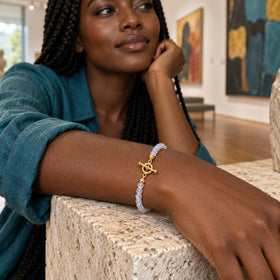 A yellow gold toggle clasp and pale moonstone beads encircle a woman's wrist resting on a stone console in an art gallery.