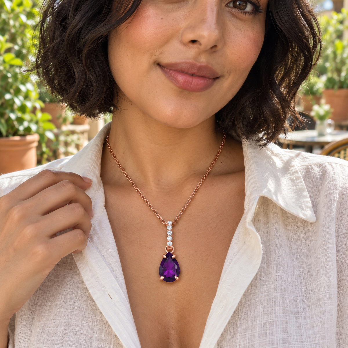 A woman in an open white linen shirt wears a rose gold chain with a purple pear-shaped pendant on a leafy cafe terrace.