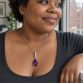 A woman in a charcoal top wears a silver chain with a purple amethyst pendant beside a window overlooking brick buildings.