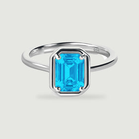 A polished silver ring with a bright blue rectangular stone sits centered on a pale grey surface.