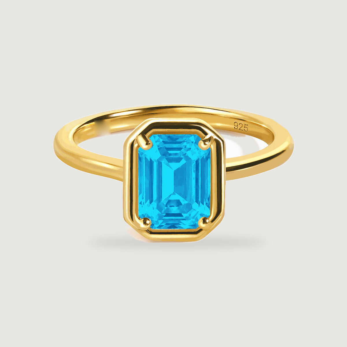 A yellow gold ring holds a blue emerald-cut stone above a soft shadow on pale grey.