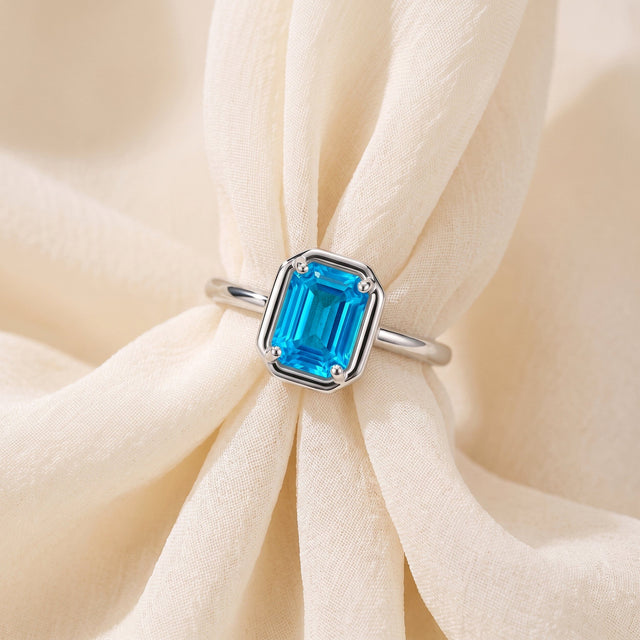 A silver ring with a vivid blue stone rests across softly folded pale fabric.