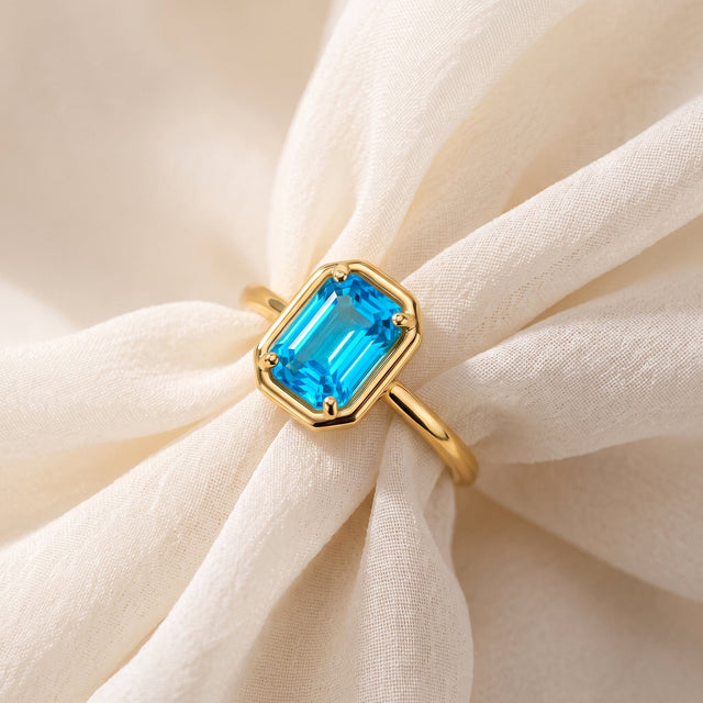 A yellow gold ring with a rectangular blue stone lies diagonally over light fabric folds.