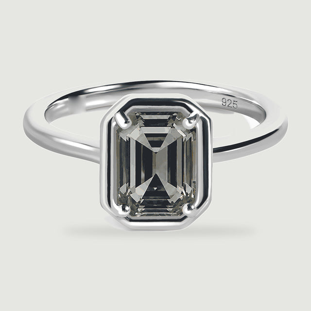 A grey emerald-cut stone shows dark stepped facets within a polished silver ring.