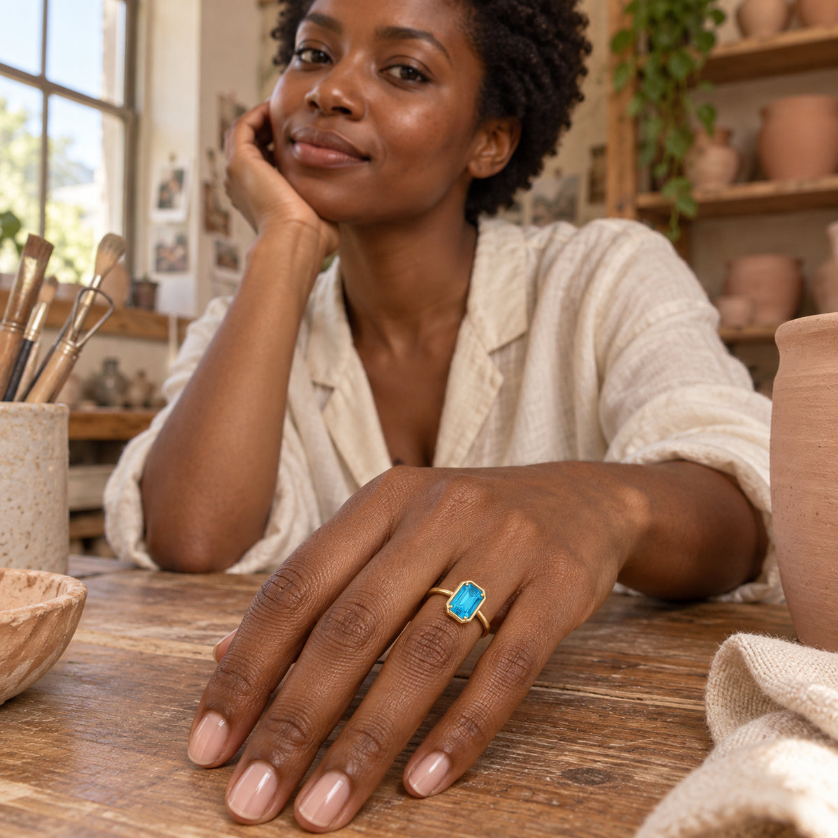 A woman in a pale linen blouse wears a blue-stone yellow gold ring with her hand resting on a pottery studio table.