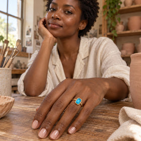 A woman in a pale linen blouse wears a blue-stone yellow gold ring with her hand resting on a pottery studio table.