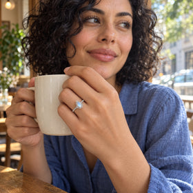 A woman in a blue linen shirt wears the white gold moonstone ring while holding a cream ceramic mug by a cafe window.