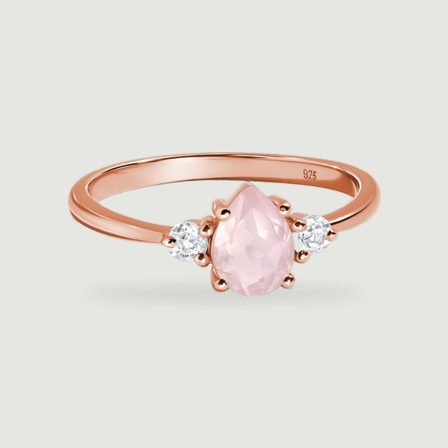 A rose gold ring with a pear-shaped pink gemstone and two round clear side stones rests on a light gray surface.