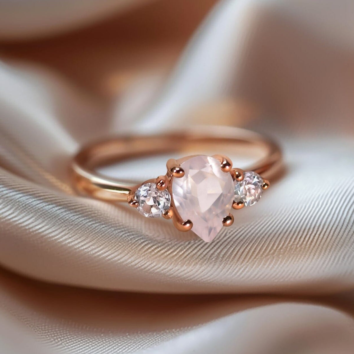 A rose gold ring with a pale pink pear-shaped stone lies on softly folded neutral fabric.