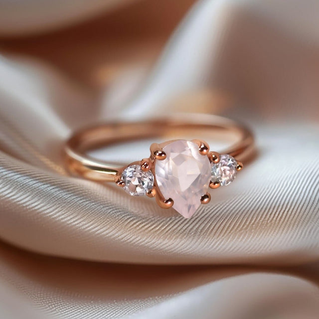 A rose gold ring with a pale pink pear-shaped stone lies on softly folded neutral fabric.