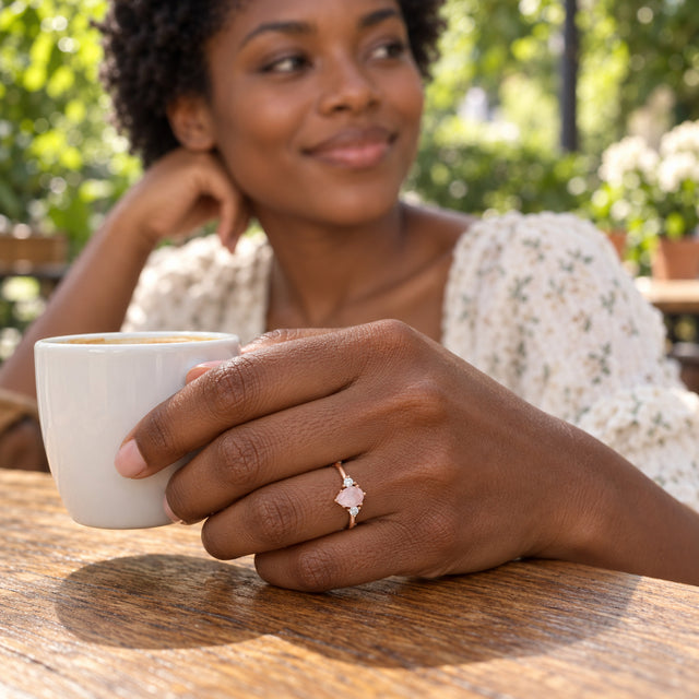 A woman with short curly hair wears the rose gold pink gemstone ring while holding a white cup at a leafy outdoor cafe.