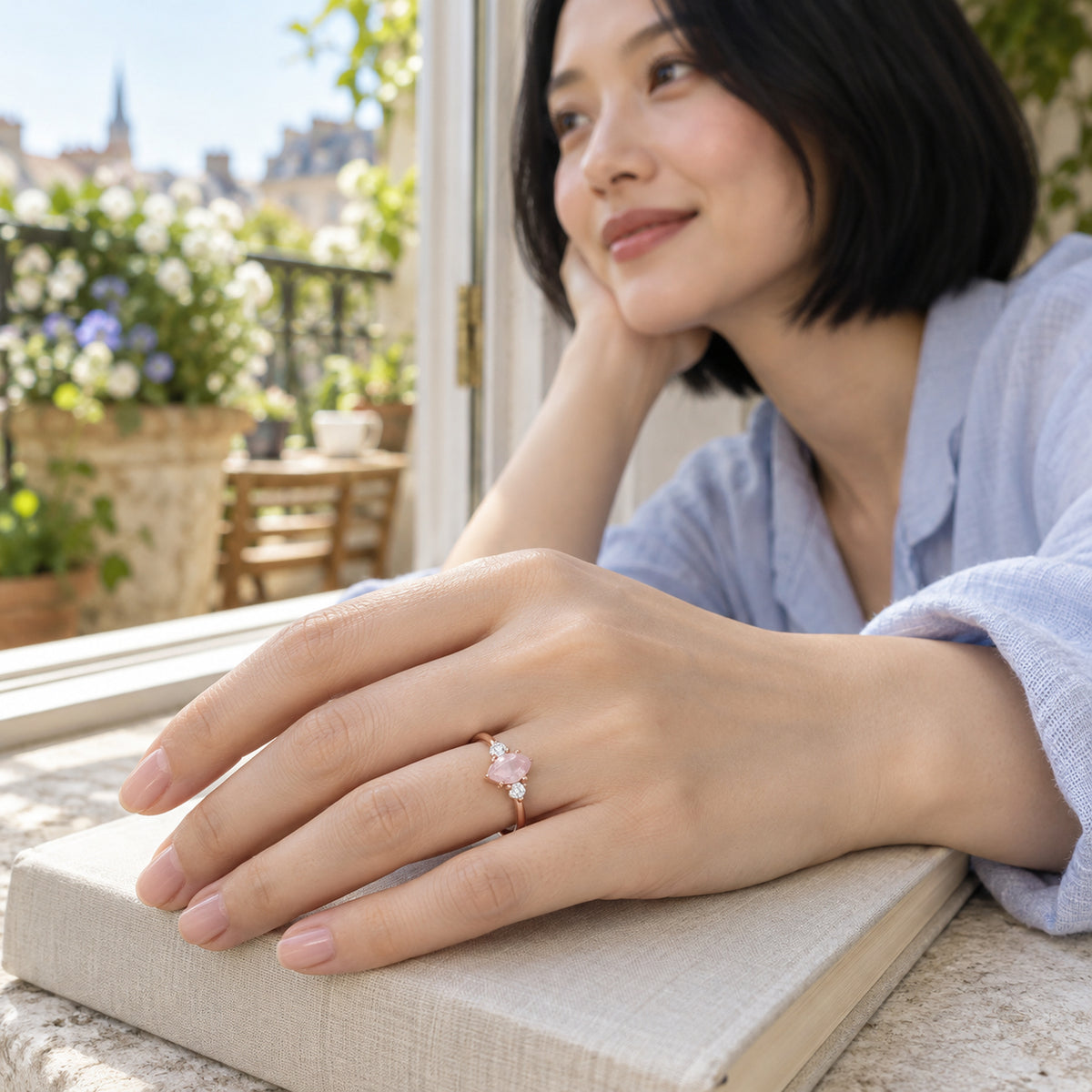 A woman with a black bob wears the rose gold pink gemstone ring with her hand resting on a linen-covered book beside a balcony window.