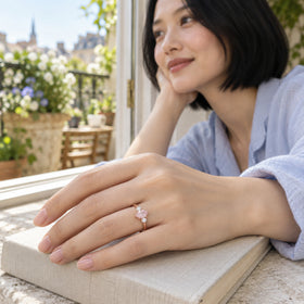 A woman with a black bob wears the rose gold pink gemstone ring with her hand resting on a linen-covered book beside a balcony window.