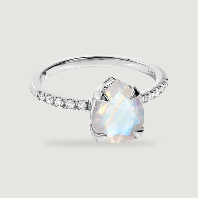 A sterling silver ring with a pear-shaped moonstone and clear stones along the band rests on a pale grey surface.