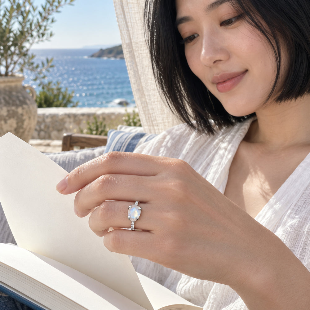 A woman with a black bob wears the silver moonstone ring while holding an open book beside a sea-facing window.
