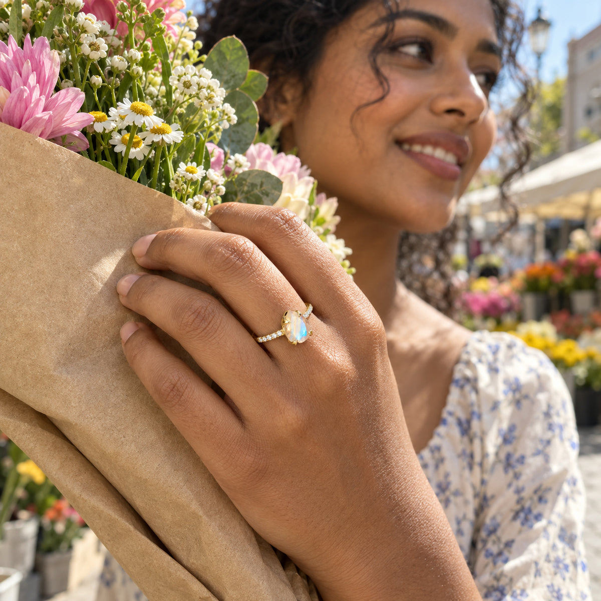 A yellow gold vermeil moonstone ring is visible on a hand holding a paper-wrapped flower bouquet at an outdoor market.