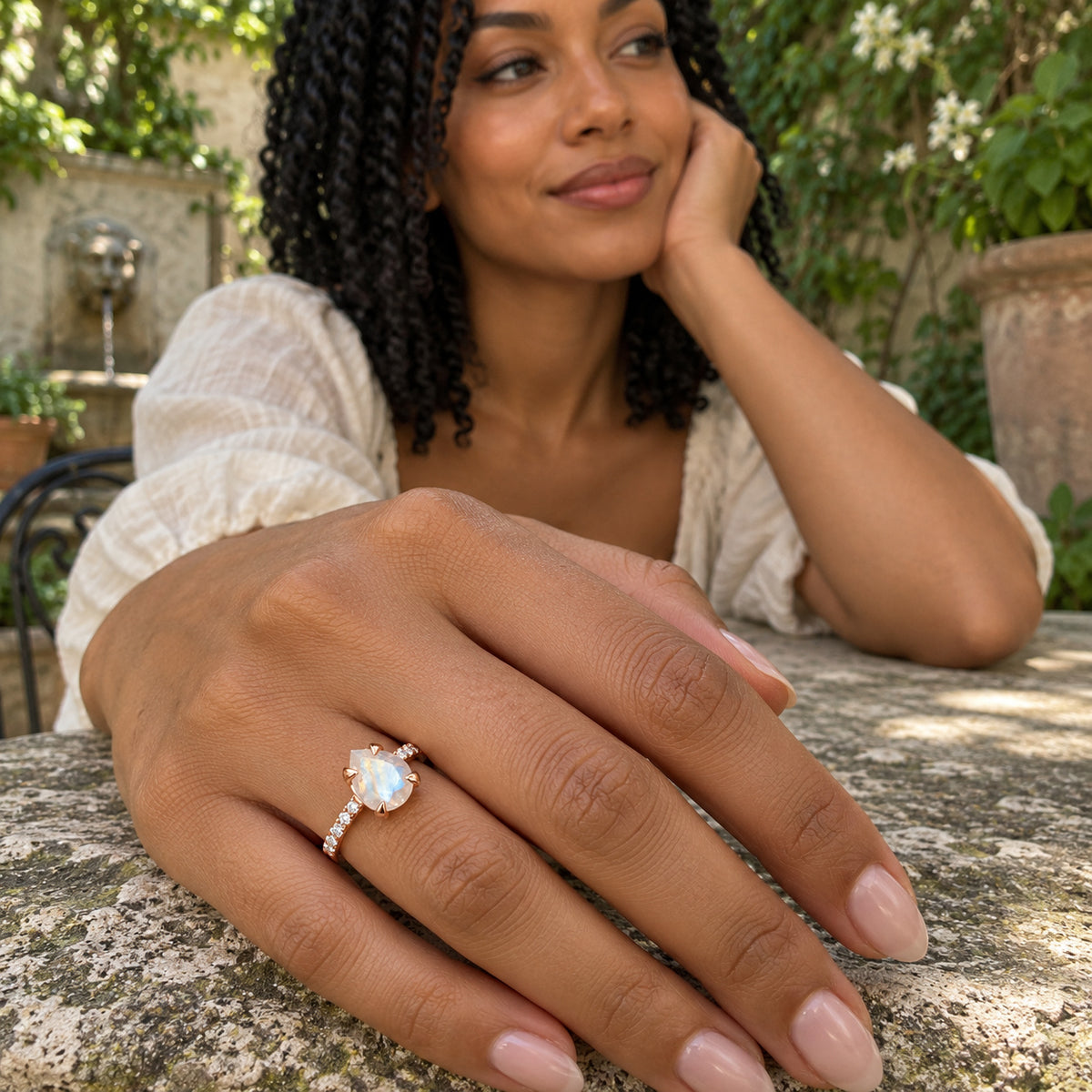 A woman with dark twists wears the rose gold vermeil moonstone ring as her hand rests on a stone table in a garden courtyard.