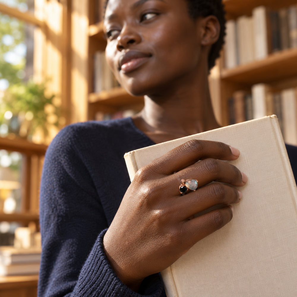 A woman with short natural hair wears the two-stone rose gold ring while holding a cream book in a sunlit bookshop.