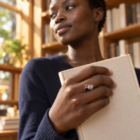 A woman with short natural hair wears the two-stone rose gold ring while holding a cream book in a sunlit bookshop.
