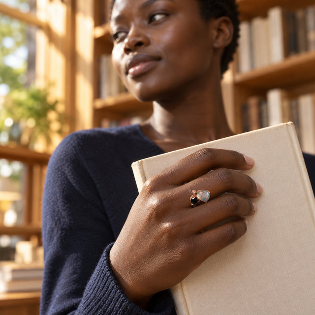 A woman with short natural hair wears the two-stone rose gold ring while holding a cream book in a sunlit bookshop.