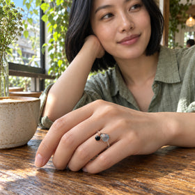 A woman with a black bob wears the labradorite and black obsidian ring with her hand resting beside a ceramic cup on a cafe table.
