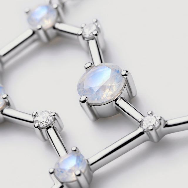An angled close-up reveals the facets of a pale moonstone and smooth silver bars beside small clear stones.
