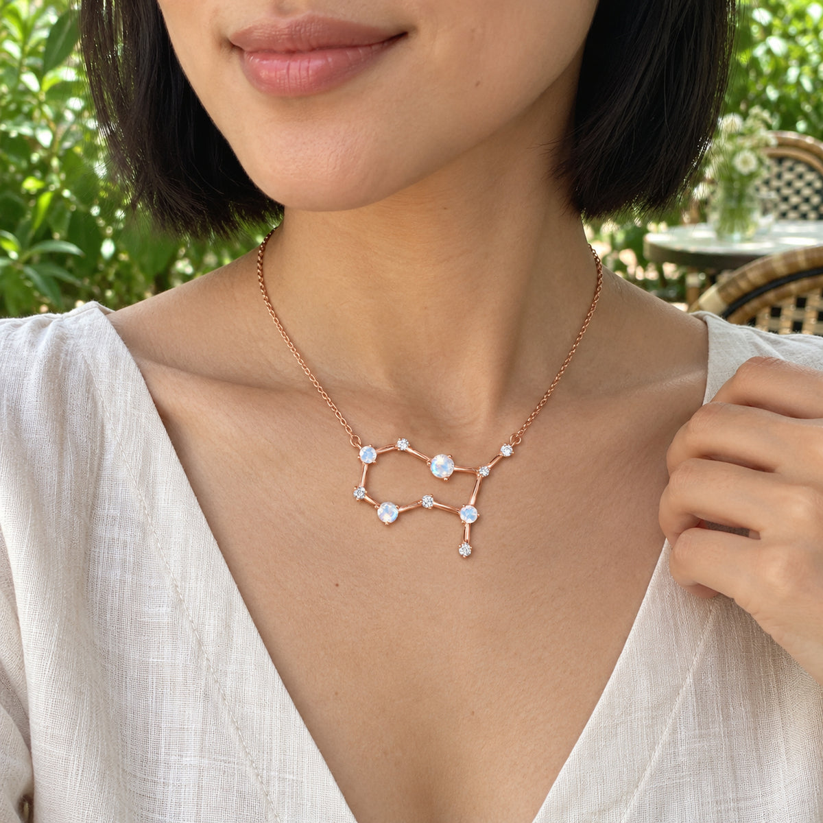 A woman wearing an ivory linen blouse displays a rose gold moonstone necklace at a leafy garden cafe.