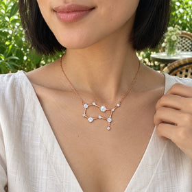 A woman wearing an ivory linen blouse displays a rose gold moonstone necklace at a leafy garden cafe.
