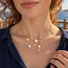 A yellow gold moonstone necklace rests below the collarbones of a woman in a navy blouse on a seaside terrace.