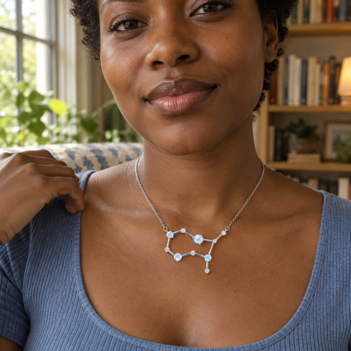 A silver moonstone necklace with an open geometric pendant is worn with a blue scoop-neck top in a book-lined room.