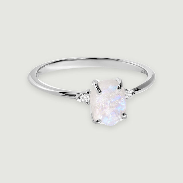 A polished silver ring with a raw pale moonstone and two round white zircon accents rests on a light grey background.