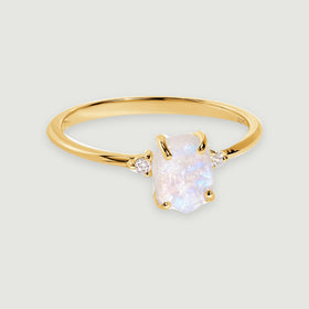 A yellow gold vermeil ring displays a rough moonstone between two small white zircon stones on a softly shadowed grey surface.
