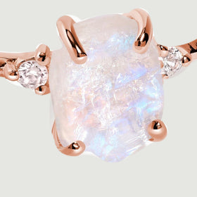 Rose gold vermeil claws frame the raw moonstone beside two sparkling white zircon accents against a light grey backdrop.