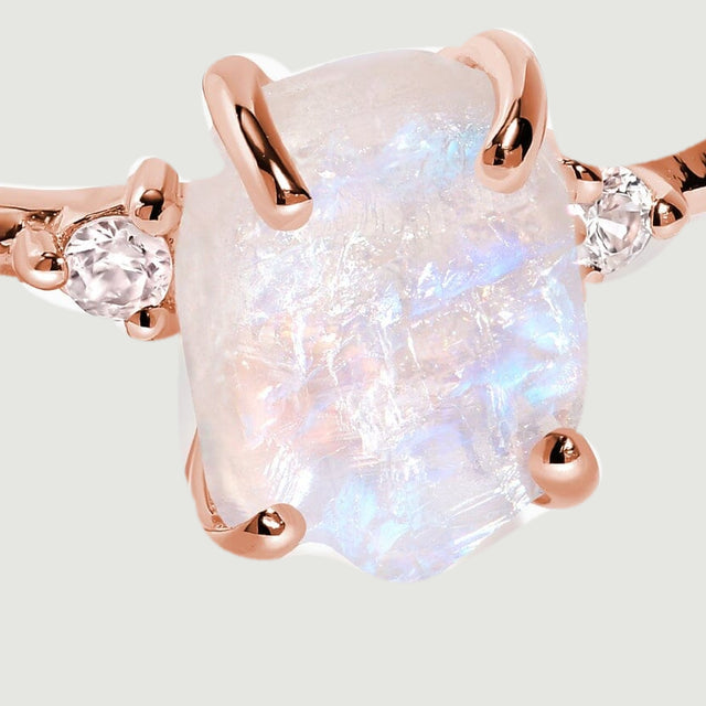 Rose gold vermeil claws frame the raw moonstone beside two sparkling white zircon accents against a light grey backdrop.