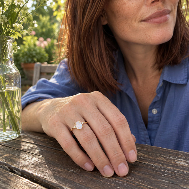 A woman in a blue shirt wears the yellow gold vermeil moonstone ring as her hand rests on a wooden patio table beside a glass vase.