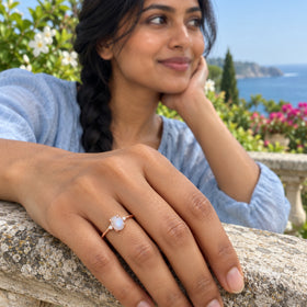 The rose gold vermeil moonstone ring sits on a woman's finger as her hand rests on a stone railing in a coastal garden.